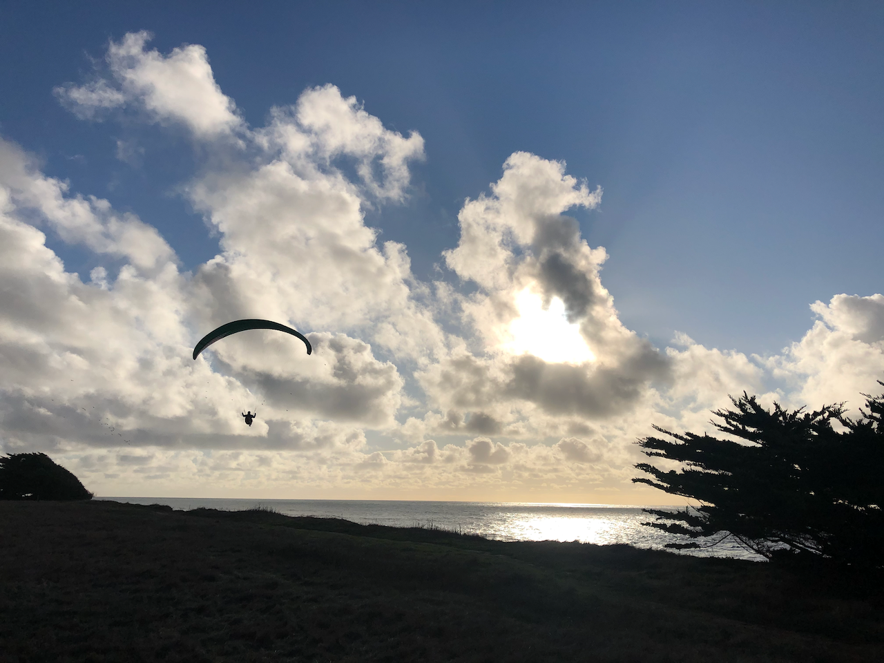 The silhouette of a paraglider floats above the bluffs as the sun descends towards the horizon, shining through the clouds and over the ocean.